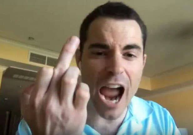 A man in a light blue shirt shouting with his mouth open and pointing his middle finger aggressively at the camera.