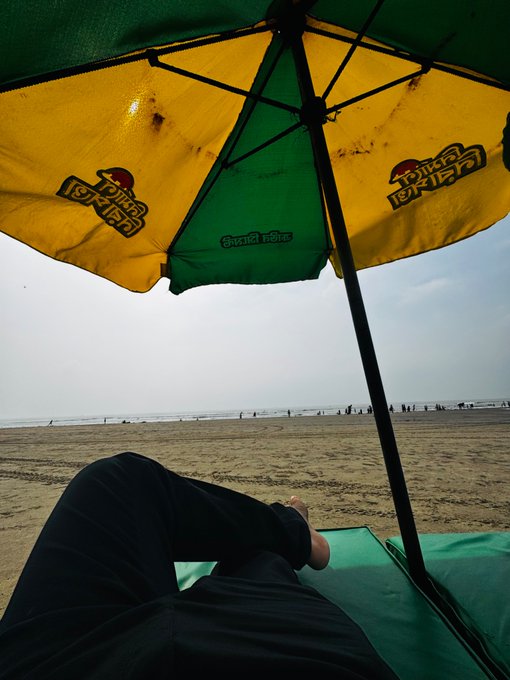 A first-person view of someone relaxing under a green and yellow 'Mirinda' (Frooti) umbrella on a beach, looking out at the ocean.