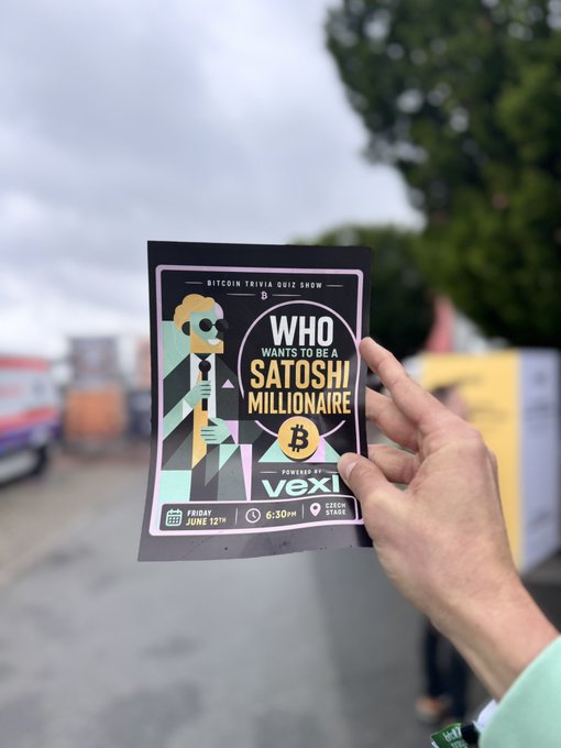 The image features a person holding up a black flyer with white text that says "Who wants to be Satoshi Millionaire? VEXI". The flyer also includes an illustration of a man holding a Bitcoin coin, which suggests that the event is related to cryptocurrency or Bitcoin. The person's hand holding the flyer appears to be in front of a truck and a building, indicating that this might be taking place at an outdoor venue or a public space.