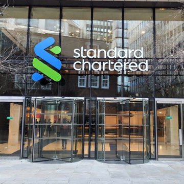 The exterior entrance of a Standard Chartered bank building featuring the company's blue and green logo on a glass facade with revolving doors.