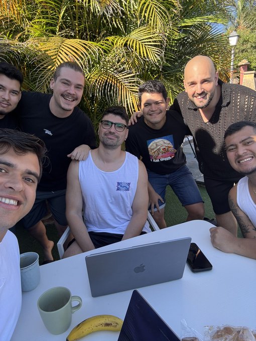 A casual outdoor group selfie of seven smiling men gathered around a table with a MacBook laptop, in a tropical setting with palm trees visible.