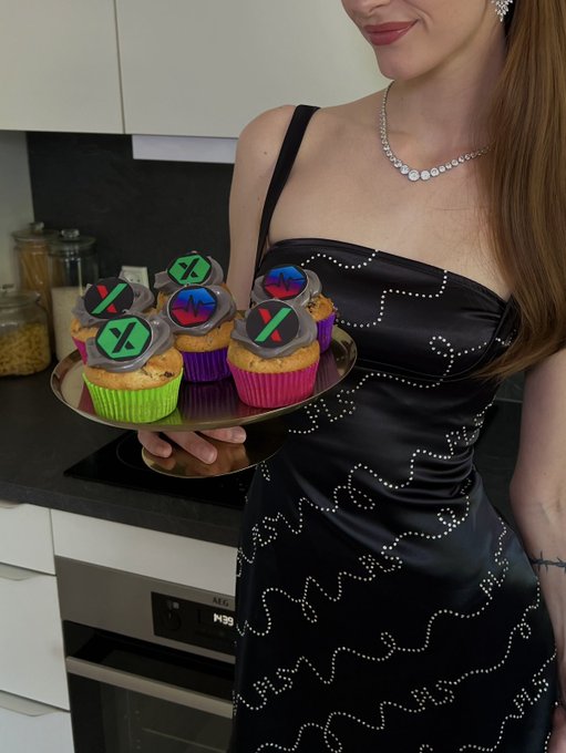 A woman in an elegant black dress with pearl embellishments and a diamond necklace holds a tray of cupcakes decorated with colorful X-shaped fondant toppers.