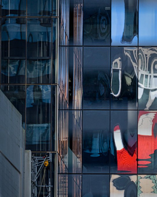 Close-up photograph of a modern glass building facade with distorted reflections of surrounding structures and colorful signage.
