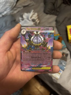 A hand holds a holographic Pokémon ex card featuring 'Phantom Manue' (130 damage) against a blurry background of bedding and other cards.