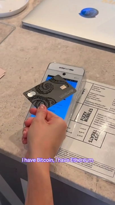 A person holds up a black "CRYPTOLINK" credit card next to an OrderPad Pro payment terminal at a cafe, with menu items like "Fizzy Bubbles" and "Slyvah Açaí" visible. The image illustrates the real-world use of the Kraken Card for paying at merchants with cryptocurrency holdings, as the caption text reads "I have Bitcoin, I have Ethereum" — suggesting the card is funded with these digital assets.