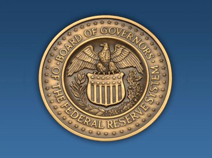 The official bronze seal of the Board of Governors of the Federal Reserve System, featuring an eagle with spread wings above a shield, set against a blue background.