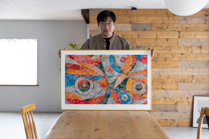 A man wearing glasses stands indoors holding a large, colorful abstract framed artwork above a wooden dining table.