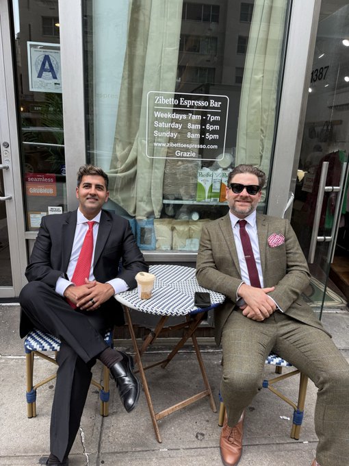 Two well-dressed men—one in a black suit with a red tie, the other in a tweed suit with sunglasses—share an espresso and conversation outside Zibetto Espresso Bar (1387), where the storefront displays hours: weekdays 7am-7pm, Saturday 8am-6pm, and Sunday 8am-5pm, suggesting a relaxed business meeting that aligns with the post's hint of "big things coming" for @solanaspaces.