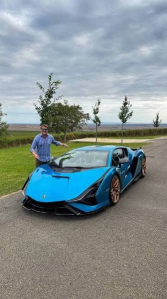 A man standing next to a bright blue Lamborghini sports car parked on an asphalt path with a cloudy sky and trees in the background.