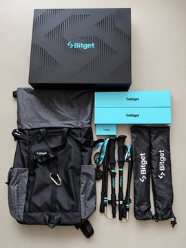 A collection of Bitget merchandise laid out, including a black backpack, black drawstring bag with the Bitget name, teal gift boxes, and two umbrellas, all branded with the Bitget logo.