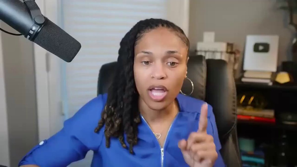 This image shows a woman sitting behind a microphone, giving an opinion or talking about something she's passionate about. She is dressed casually in blue clothes and has long dreadlocks covering her shoulders. The setting appears to be indoors, possibly a conference room, as evidenced by the presence of chairs and various items like books on shelves nearby. In the image, there are also two other people visible - one person sitting further back from the woman with the microphone and another person partially in the foreground.