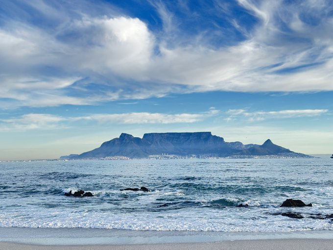 A scenic view of Table Mountain in Cape Town, South Africa, as seen from across the ocean with a sandy beach and rocky shoreline in the foreground.
