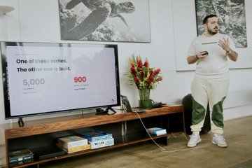 A man in a white t-shirt and sweatpants presents beside a TV screen displaying 'One of these scales. The other one is butt.' with statistics showing 5,000 vs 900.