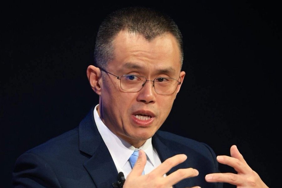 A photograph of a man with short hair and glasses wearing a dark blue suit and light blue tie, gesturing with both hands as if speaking during a presentation.