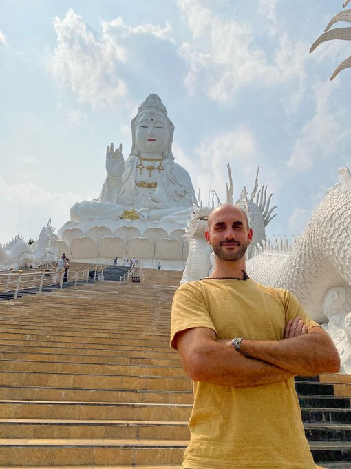 A man in a yellow t-shirt stands with crossed arms in front of a large white seated Buddha statue, likely at Wat Huay Pla Kang in Chiang Rai, Thailand.