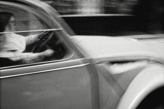 A black and white motion-blurred photograph of a woman driving a vintage car, captured with a panning technique that emphasizes the sense of speed.