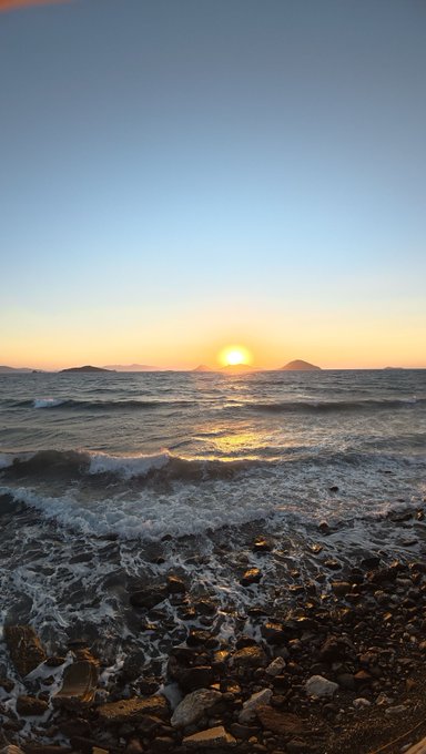 A scenic sunset over the ocean with the sun low on the horizon, casting golden light on waves crashing against a rocky pebbled shore.