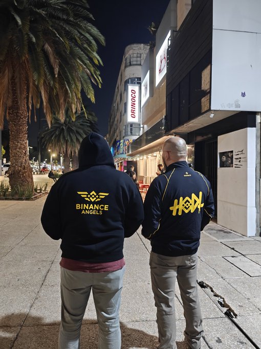 Two people standing on a city street at night, wearing hoodies; the left person wears a black 'Binance Angels' hoodie and the right person wears a blue jacket with yellow 'Hexa' lettering.