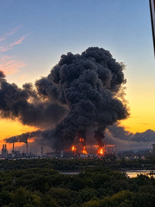 A dramatic photograph of a massive fire at an industrial facility, with a towering plume of thick black smoke rising into a sunset-colored sky.