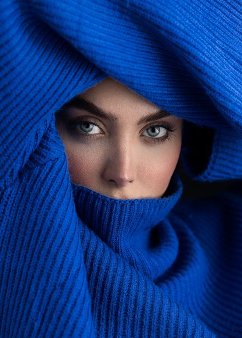 A close-up portrait of a young woman with striking blue eyes, her face partially obscured by a vibrant blue ribbed fabric wrapped around her head and neck.