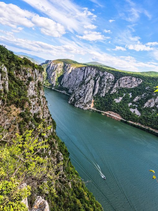 A scenic landscape photo of a river gorge with steep limestone cliffs covered in green vegetation, a boat leaving a wake on the green water, and a bright blue sky with clouds.