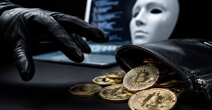 A dark conceptual image showing a gloved hand reaching toward a spilled pouch of gold Bitcoin coins, with a white mask and a laptop displaying code in the background.