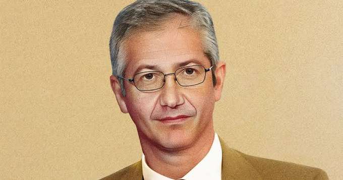 A digital illustrated portrait of an older man with gray hair and glasses wearing a tan suit jacket against a beige background.