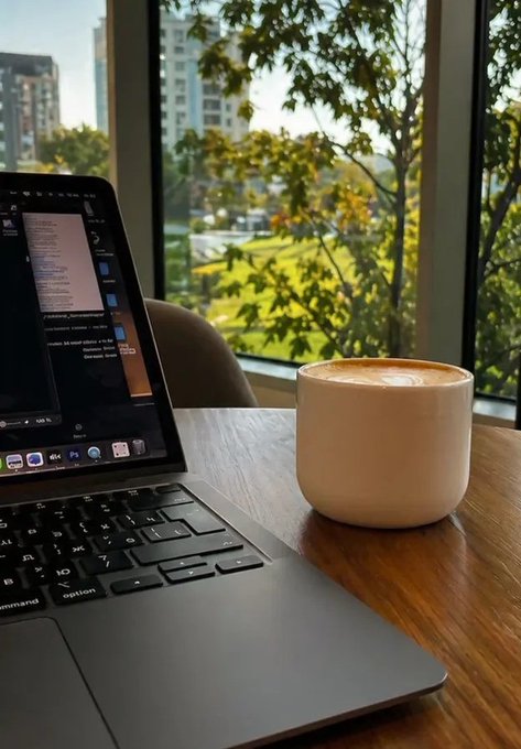 The image features a laptop computer sitting on top of a wooden table, with a coffee cup placed next to it. The scene takes place outside, possibly near a window overlooking a park or another outdoor area. A person can be seen in the background, but their face is not visible in the photo. The focus of the image seems to be on the laptop and its surroundings rather than the person present.