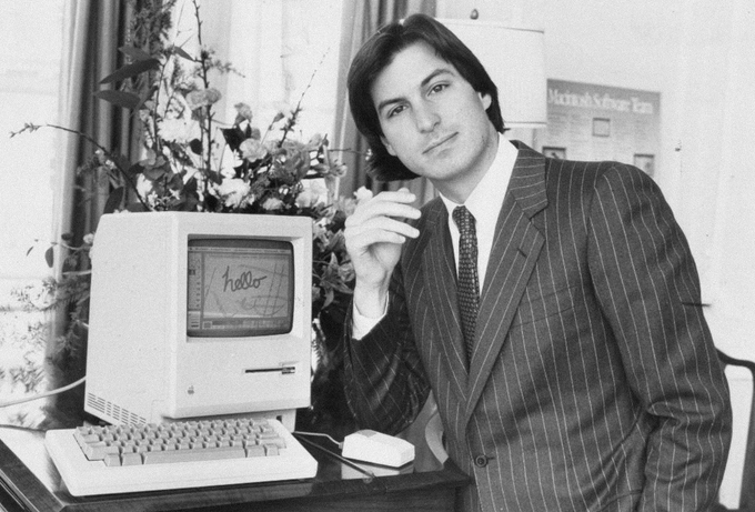 A black-and-white photograph of a young man in a pinstripe suit posing beside an early Macintosh computer displaying the word 'hello'.