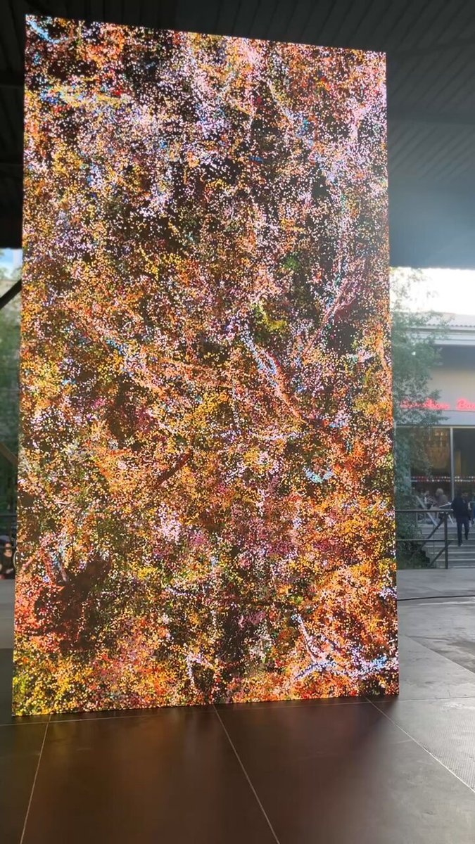 The image shows a large, colorful painting displayed outdoors. The painting features a variety of colors and patterns that create an abstract and visually striking display. In the background, there are trees visible behind the artwork, adding depth and context to the scene. The painting is situated on a tiled floor, which contrasts with its vibrant colors and enhances its visual impact. The post accompanying the image provides information about the event taking place at Basel Evening reception for @quayola, scheduled for @TezosFoundation x HEK 2026.