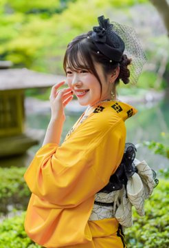 A young woman wearing a yellow kimono and a black netted hair accessory, smiling as she looks over her shoulder in an outdoor garden setting.