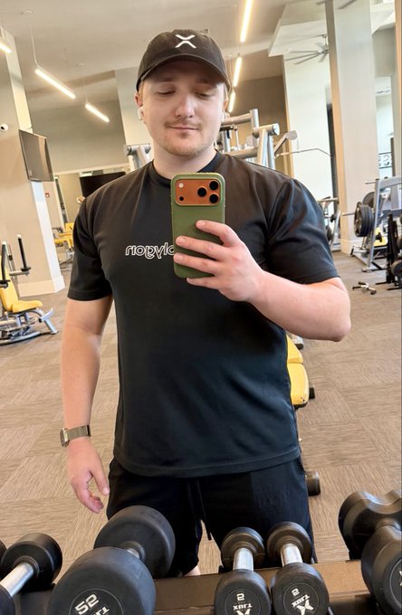 A man wearing a black cap and 'nopic' t-shirt takes a mirror selfie in a gym, with dumbbells and workout equipment visible around him.