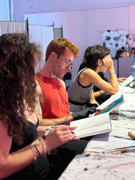 A photograph of people seated at a table, including a man in a red shirt with glasses, appearing to take notes or read materials during an event or workshop.