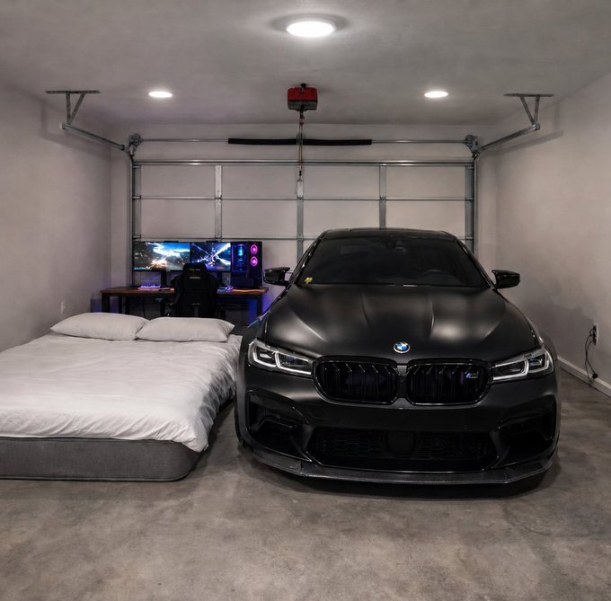 A garage interior featuring a matte black BMW sedan parked next to a bed, with a gaming/computer setup visible in the background under recessed lighting.