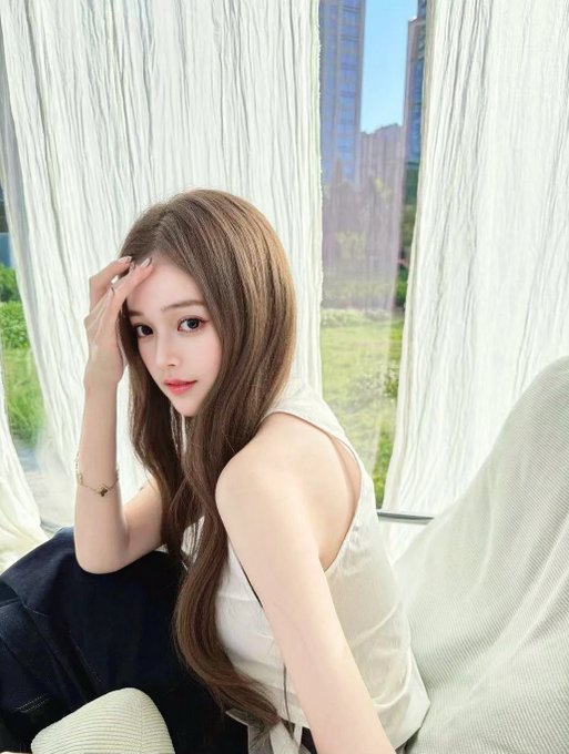 A young woman with long brown hair sits on a couch in front of a window with sheer white curtains, wearing a sleeveless white top and dark jeans.