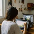 The image shows a woman sitting at a desk with two laptops, one of which has an open drawing program that she is using. The other laptop appears to be a desktop computer. There are several pictures hanging on the wall behind her, adding a personal touch to the space. The woman's back is turned towards the viewer, and there is text in the image that reads "x3 GTD spots for MEMEHAUS -> follow @memehaus_eth & @Totinhiiio - like & retweet this post - drop EVM wallet Winners in 24h".