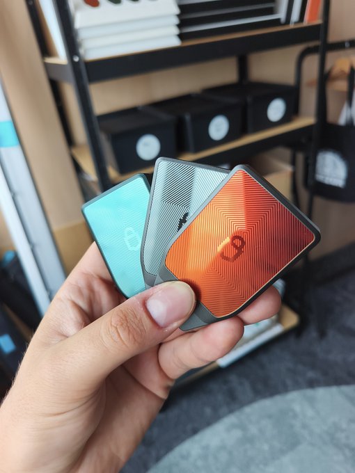 A hand holding three colorful card holders (in teal, gray, and orange) with logos, displayed in front of a retail store shelf with various accessories.