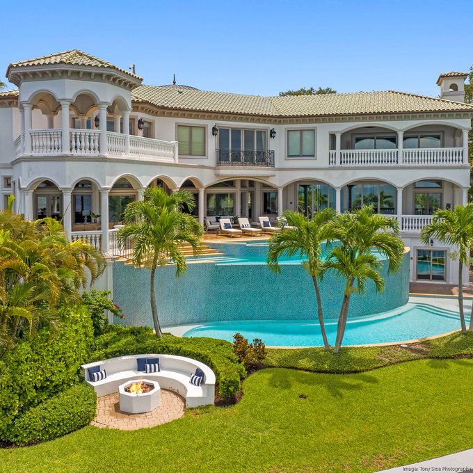 A luxurious Mediterranean-style mansion with arched windows, a curved infinity pool, palm trees, and a fire pit lounge area.