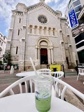 A green iced drink in a clear plastic cup sits on a white table, with a large historic stone church building visible in the background.
