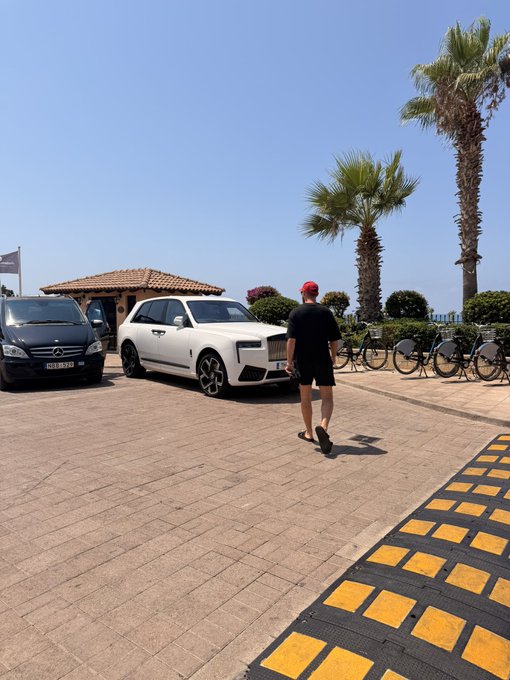 A man in a black shirt and red cap walks through a sunny coastal parking area past a white Rolls-Royce, with palm trees and bicycles lining the background. The "Gm friends" greeting pairs well with the relaxed, upscale morning vibe of the seaside scene.