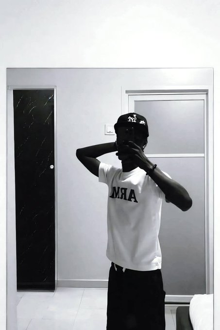 A black-and-white mirror selfie of a person wearing a 'BAMA' t-shirt, dark pants, and a cap, with their hands raised near their face.
