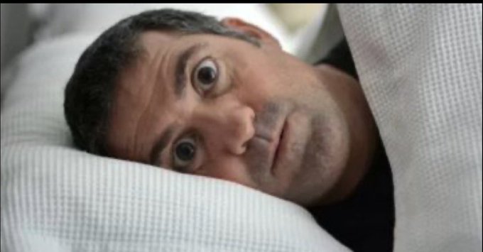 A man lying in bed wrapped in a white blanket, staring wide-eyed at the camera with a shocked or startled expression.