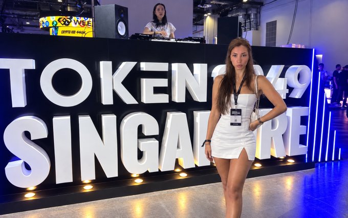 A woman in a white dress poses in front of the "TOKEN2049 SINGAPORE" signage at the crypto conference, with a DJ booth visible behind her. The image relates to the post by showing the influencer-style photo at the event, playing on the "super intelligence" joke by appearing at the Singapore conference despite the caption suggesting AI-generated presence.