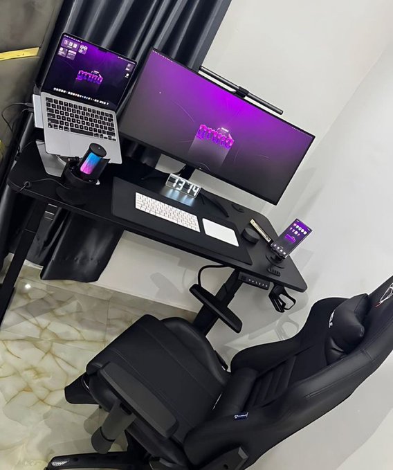A sleek black desk hosts a laptop and a large monitor, both displaying a purple 'GMAA' wallpaper, with a digital clock reading 13:49, all set within a modern room featuring a black gaming chair.