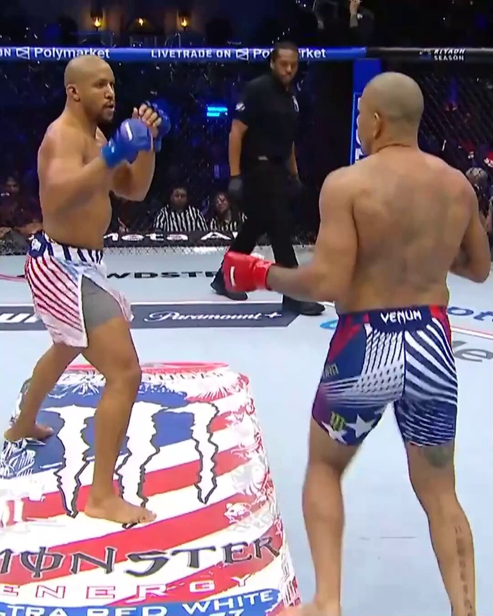 The image shows two men dressed in colorful shorts, engaged in a fight during a professional MMA match. The crowd appears to be watching intently as the fighters exchange blows. The fighter on the left has his mouth open, while the fighter on the right is holding up his fist. Both are wearing gloves and appear to be in the middle of an intense bout. The image also includes text that reads "The crowd makes their favorite known #UFCWhiteHouse LIVE NOW on @ParamountPlus [ Title fights presented by @Cryptocom ]".