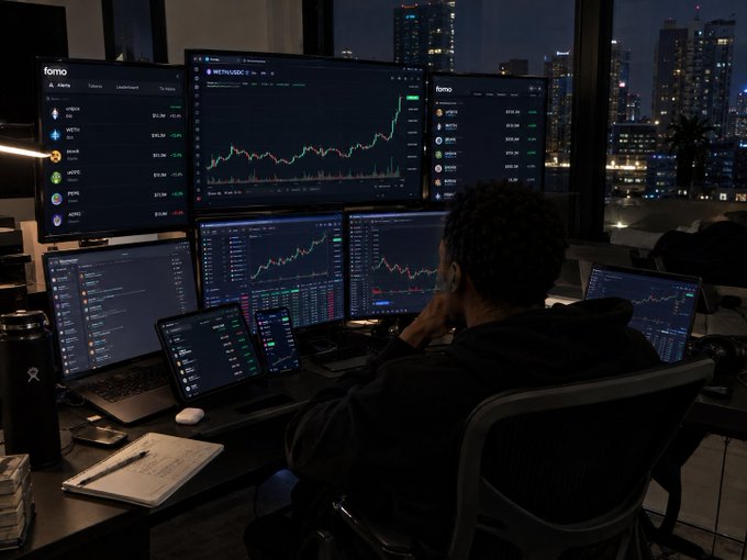 A person sits before a multi-monitor workstation at night, with screens displaying the "fomo" trading platform showing cryptocurrency tickers like PEPE, WIF, FLOKI, BONK, and DOGE alongside price charts. The image visually reinforces the post's message by depicting the kind of dedicated, screen-intensive crypto trading setup the author implies is necessary to avoid being "NGMI" (not going to make it) in the markets over the coming months.