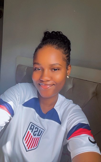 A smiling woman poses for a selfie wearing a white USA soccer jersey, seated on a tufted gray couch.