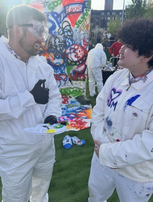 Two smiling people in white painter's coveralls and safety goggles at an outdoor event, with one holding a paint palette near a graffiti-covered backdrop reading 'Life is Gold' and 'Love'.
