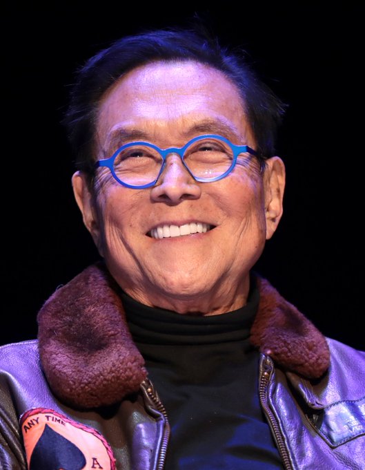 A smiling man with blue-rimmed glasses wears a brown leather jacket with a shearling collar over a black turtleneck.