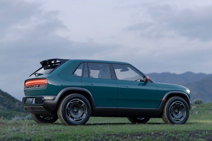 A dark green Rivian R3 electric SUV shown in a rear three-quarter view, parked on a grassy landscape with mountains in the background.